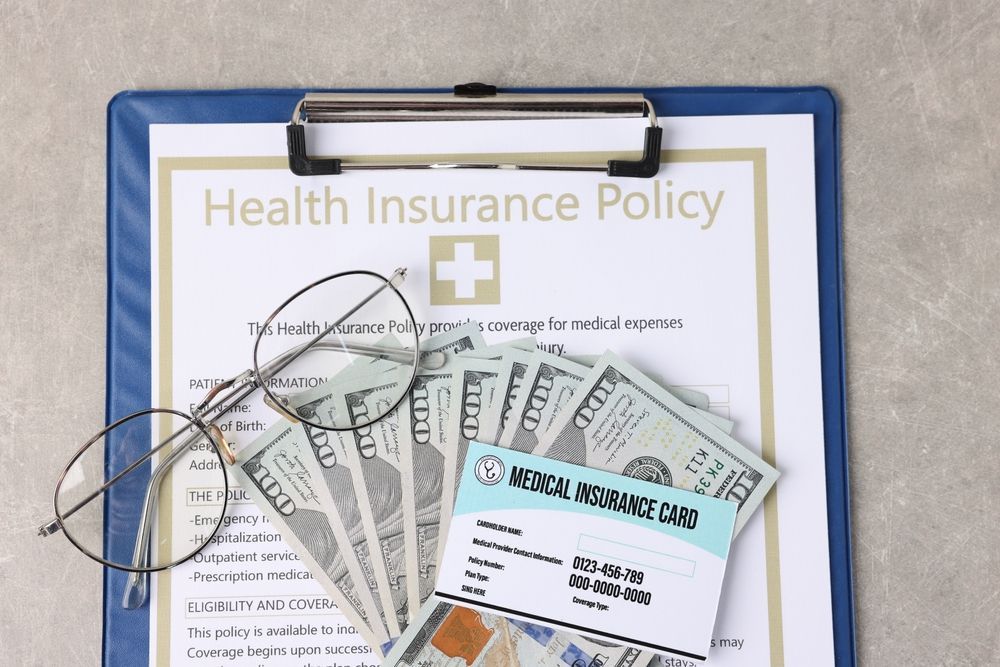Health insurance policy, money, glasses, and medical insurance card on a clipboard.