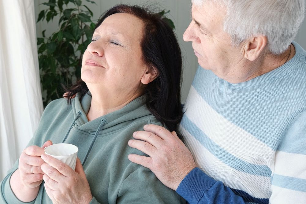 An elderly couple stands by a window. The woman holds a mug, while the man comforts her, placing a hand on her shoulder.
