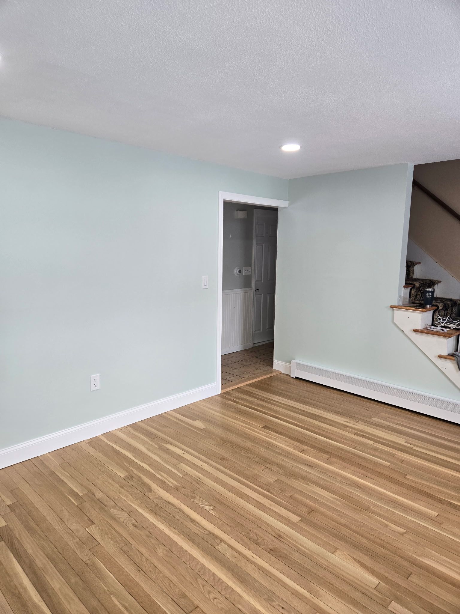 Empty room with light blue walls, wood floor, and an open doorway beside a staircase.