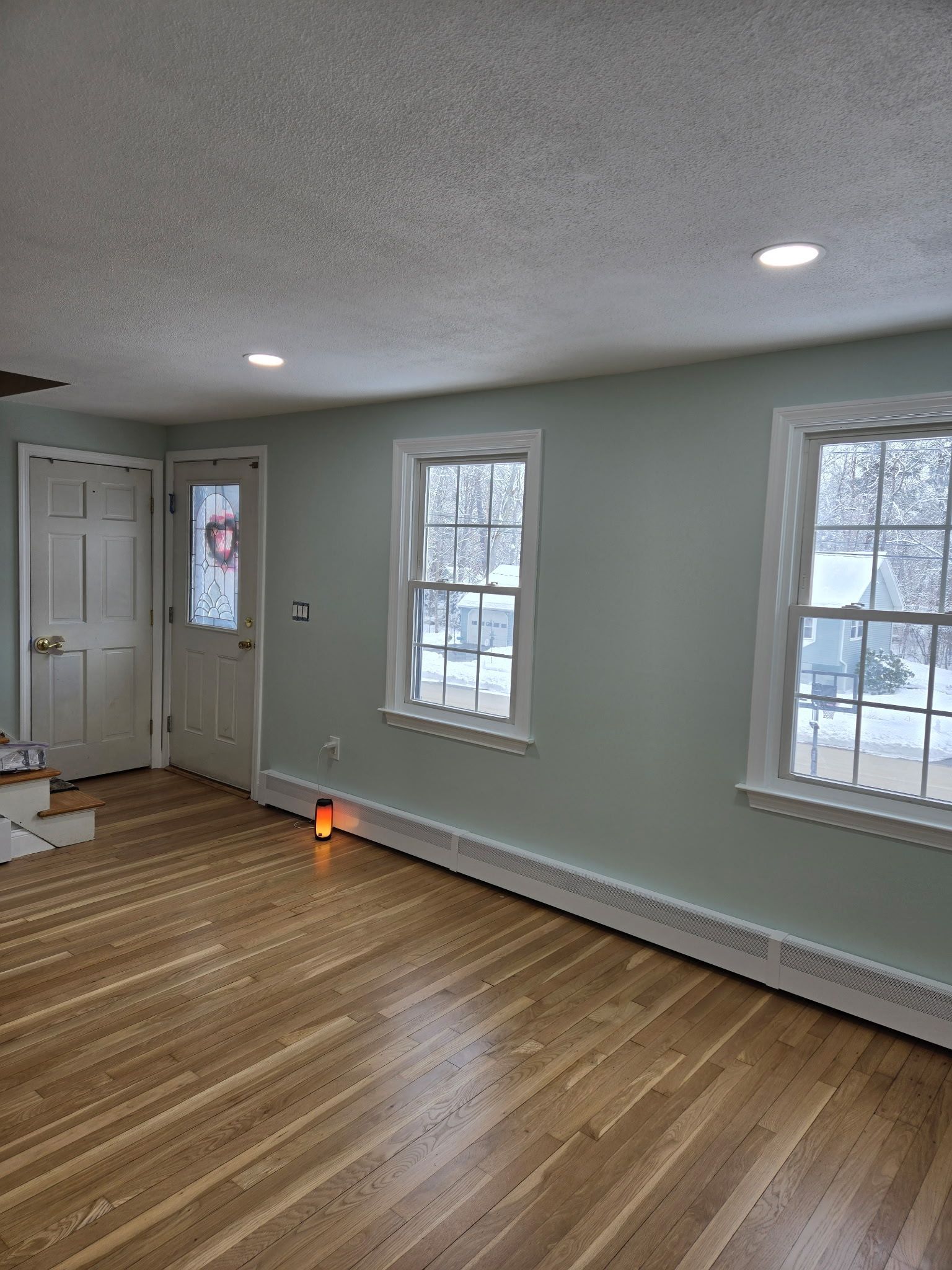Empty sunlit room with hardwood floors, pale walls, white trim, and three windows near a door.