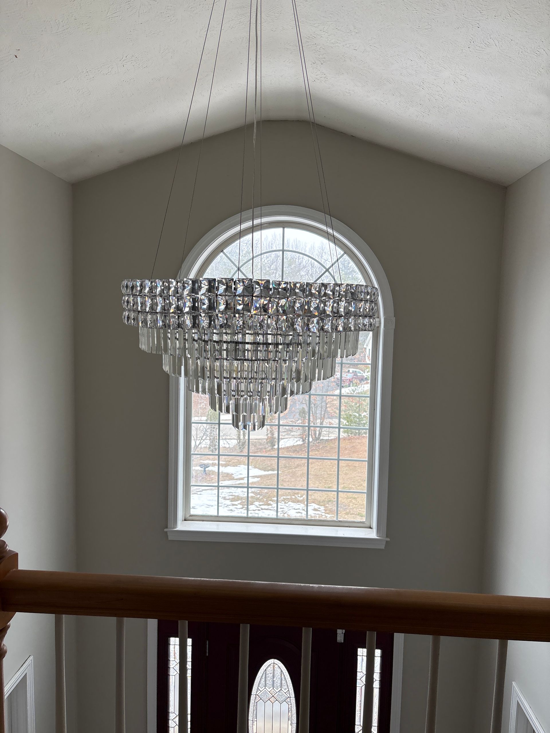 Crystal chandelier hanging in a beige stairwell beside an arched window, with a wooden railing below.
