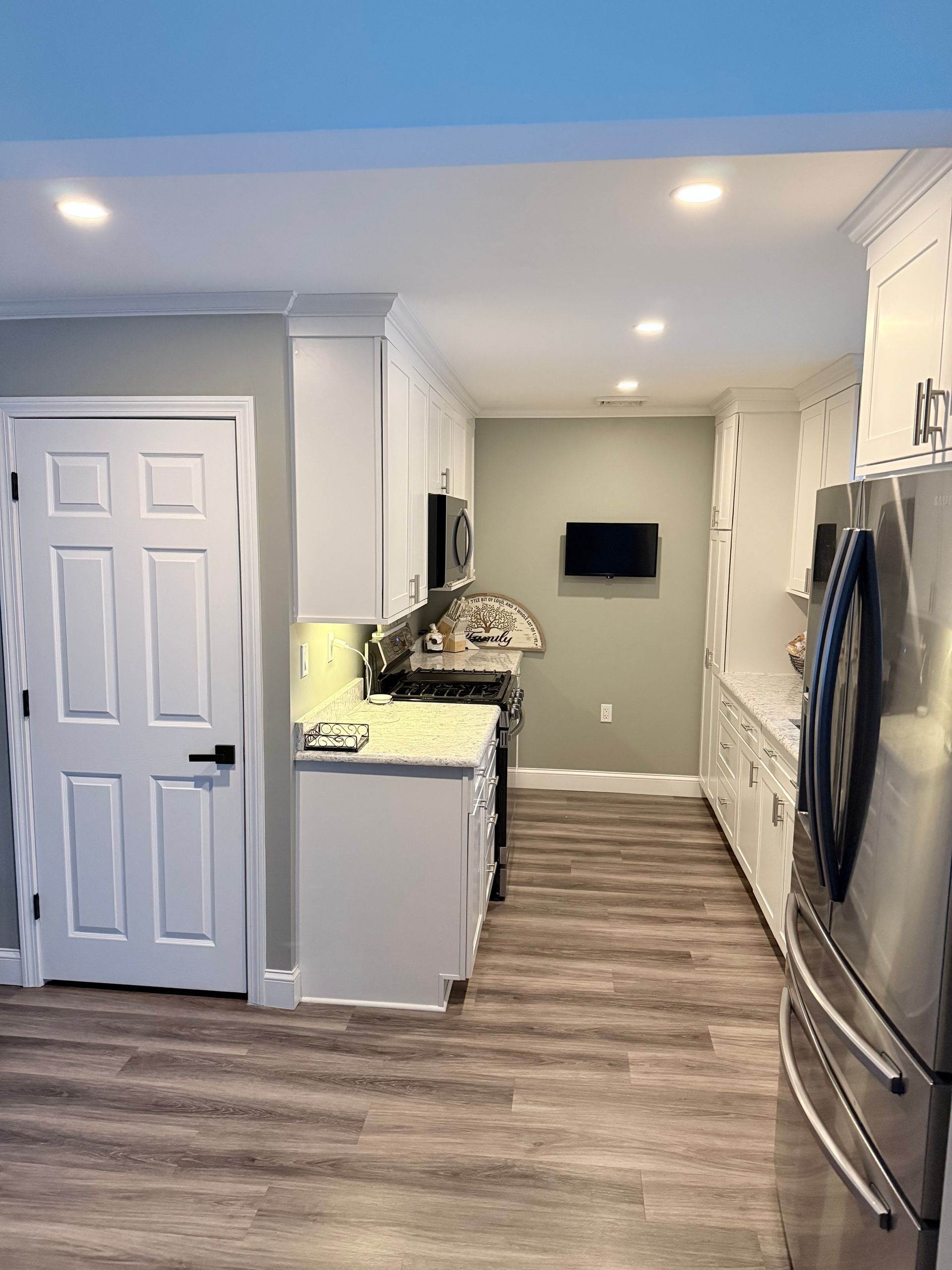Bright kitchen with white cabinets, wood-look floor, and wall-mounted TV down a narrow hallway
