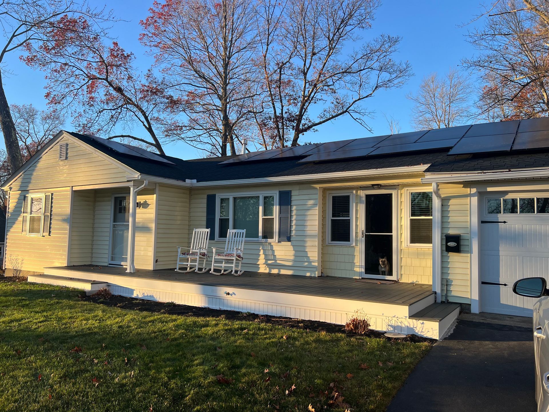 Single-story yellow house with a front porch, white chairs, and a solar-paneled roof in daylight