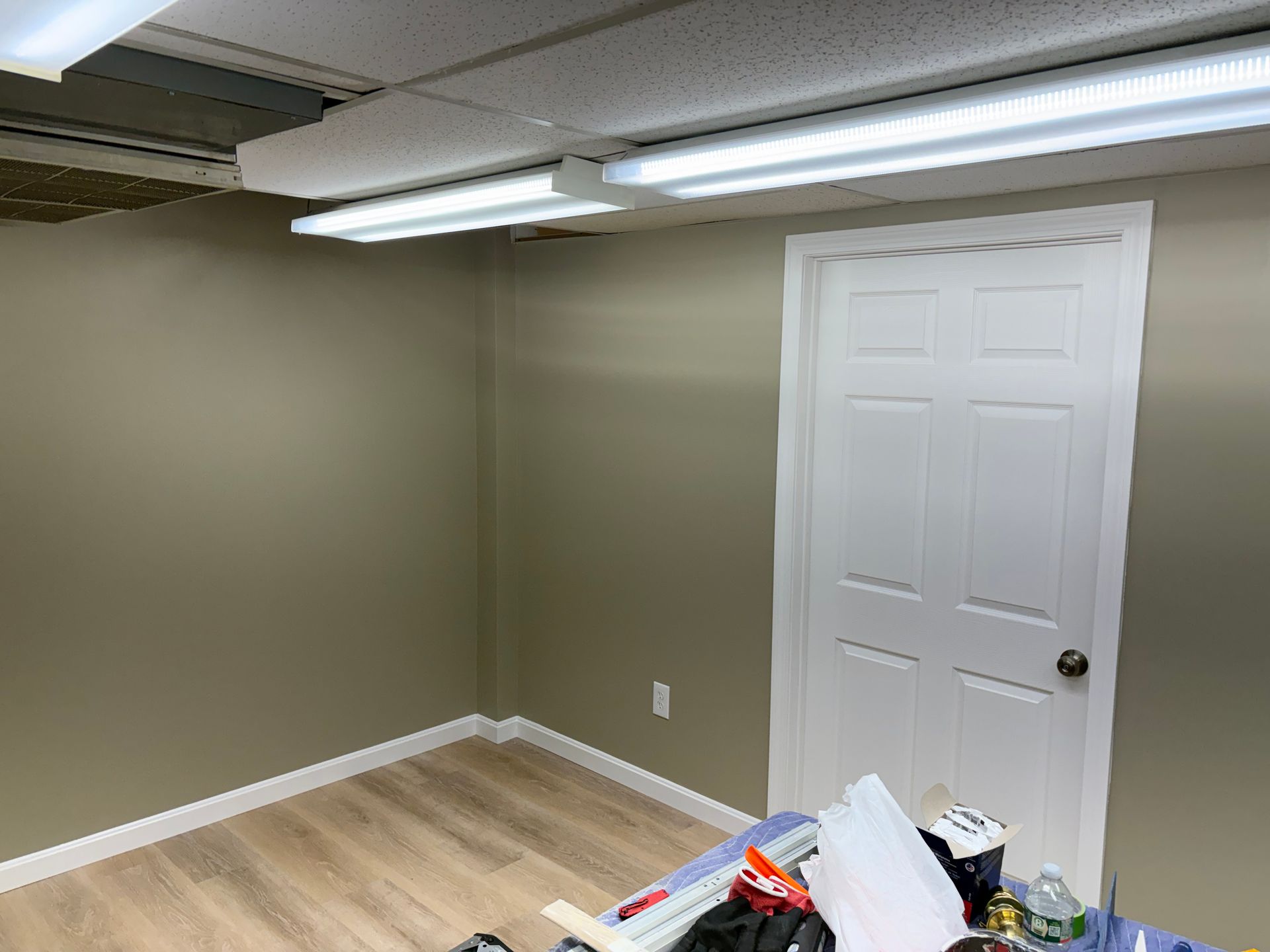 Empty room with white door, tan walls, and wood-look flooring, lit by fluorescent lights.