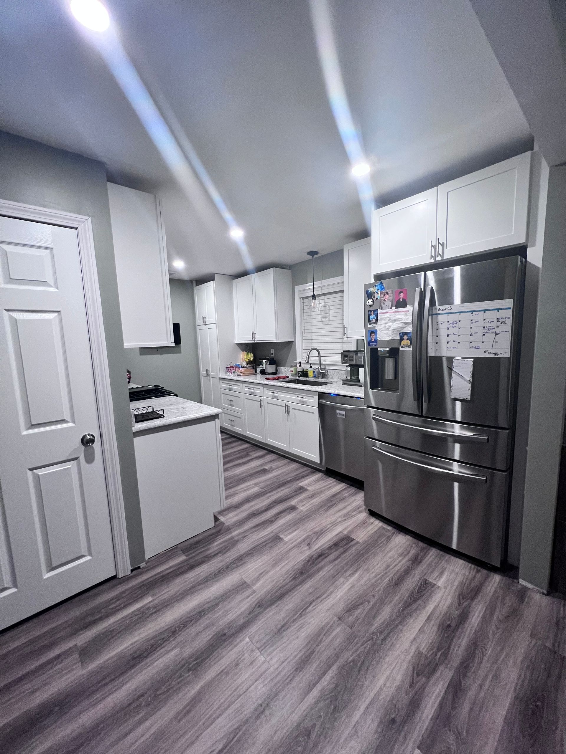 Bright white kitchen with gray wood-look floors and stainless steel appliances, viewed down a narrow hallway.