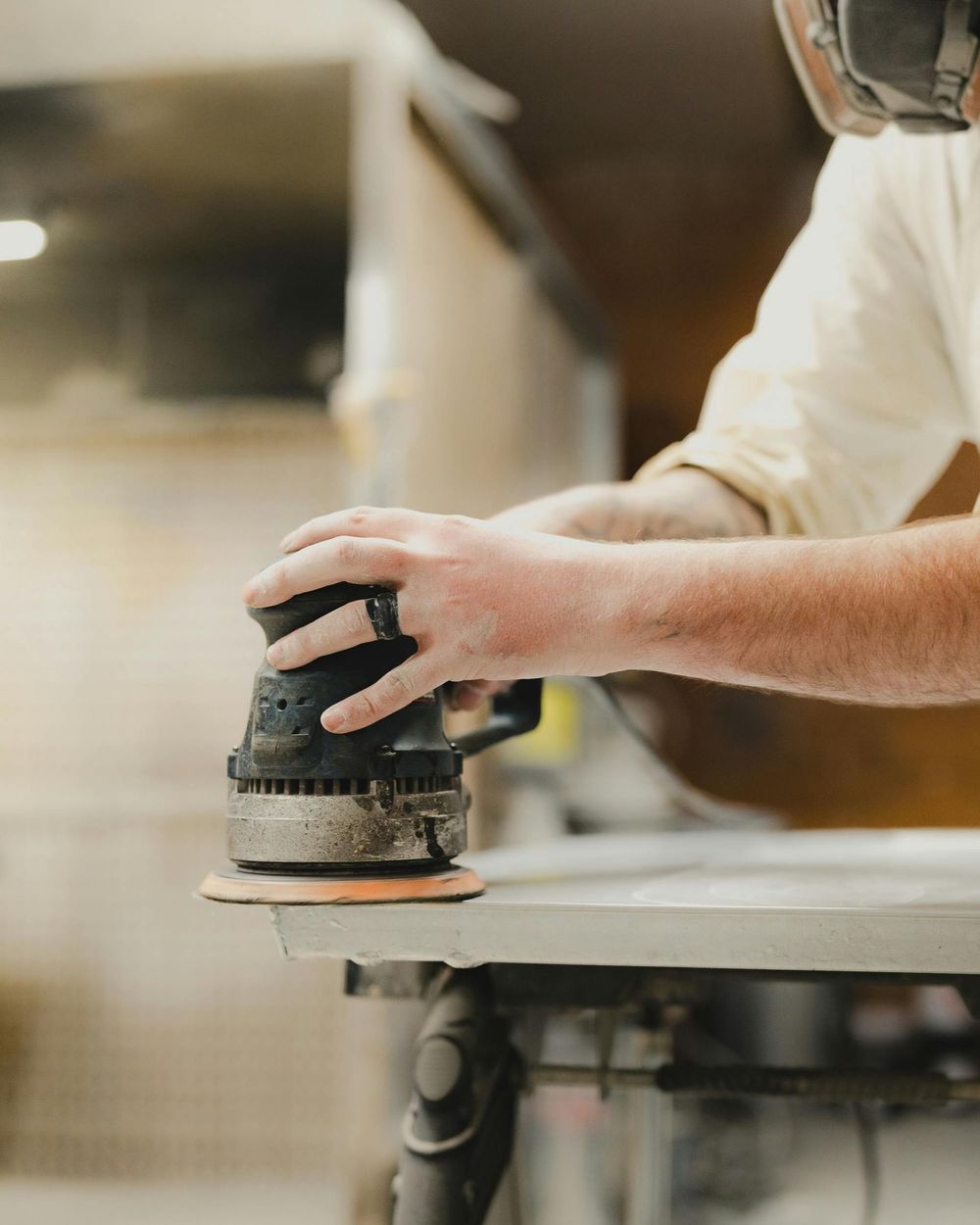 Person sanding a surface with an orbital sander in a workshop; focus on hands and tool.