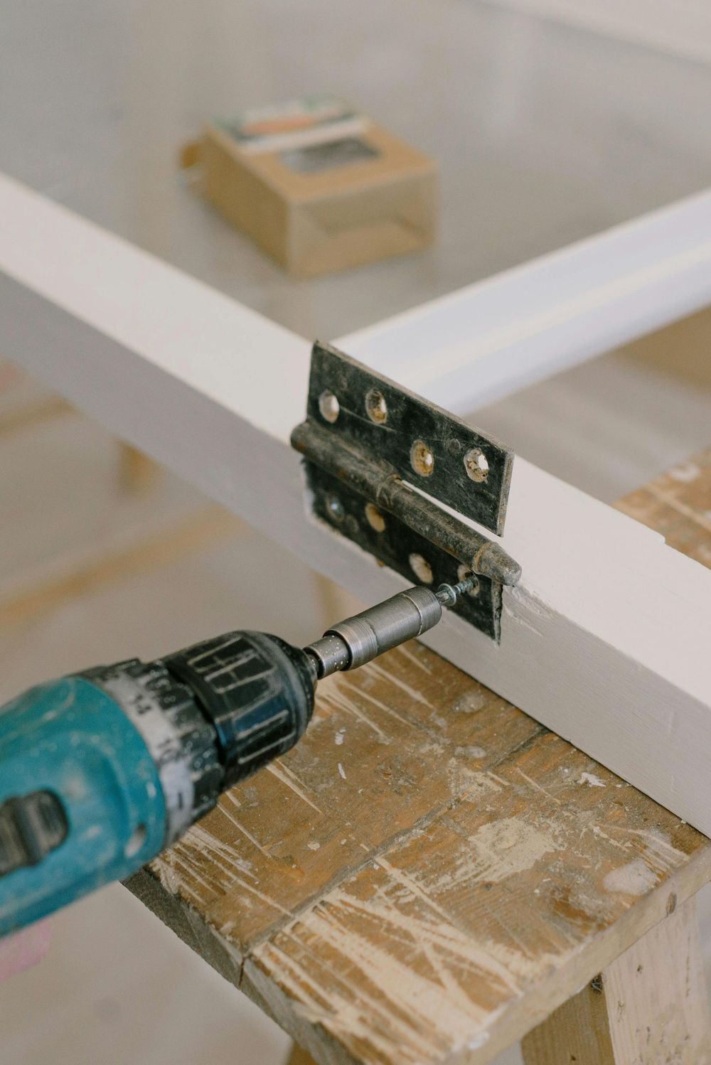 A hand using a power drill to install a hinge on a white wooden frame.