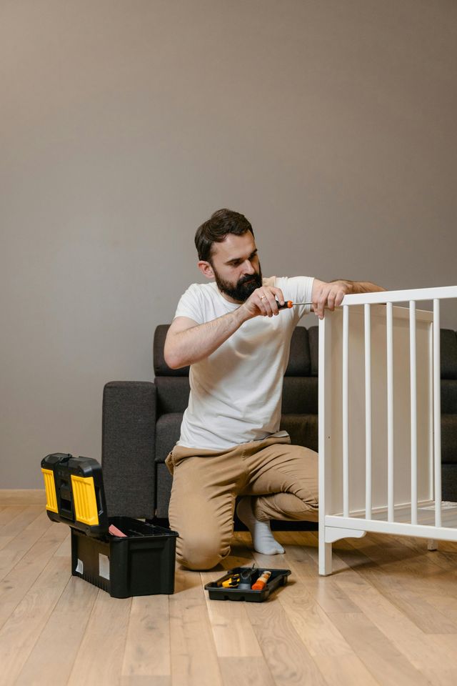 Man assembling a white crib on a wooden floor, with a toolbox nearby.