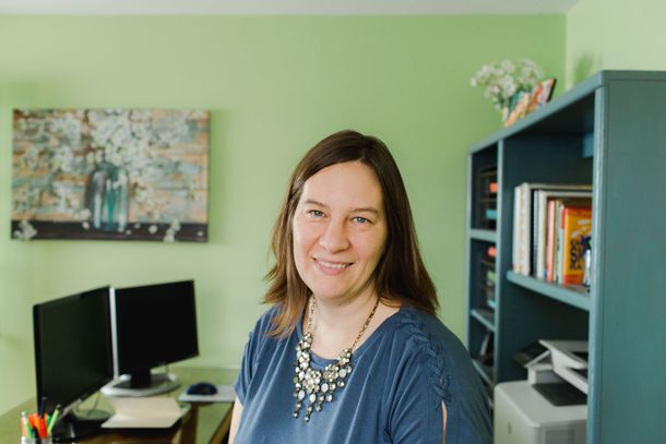 Woman smiling in home office, teal bookcase, mint green wall, desk with monitors, art.