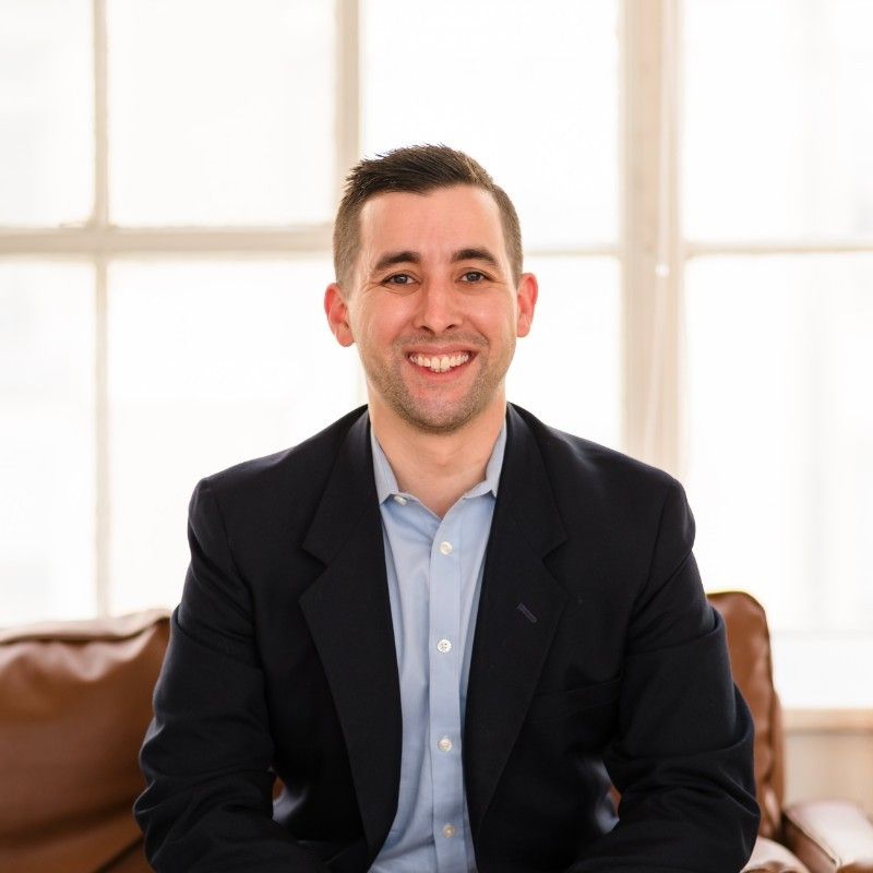 Smiling man in a dark blazer sits on a couch in a bright room by large windows