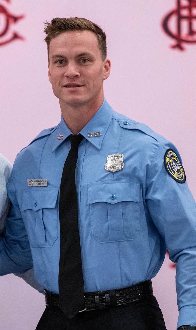A person in a light blue police uniform with a black tie and a badge, standing against a blurred background.