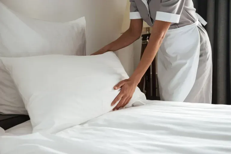 A maid is making a bed with white sheets and pillows.