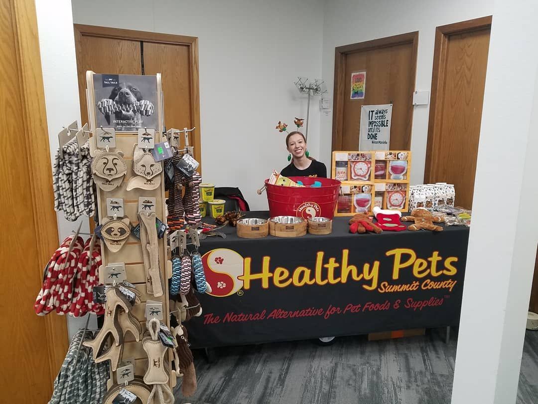 A person stands behind a Healthy Pets display table filled with pet products, treats, and accessories.