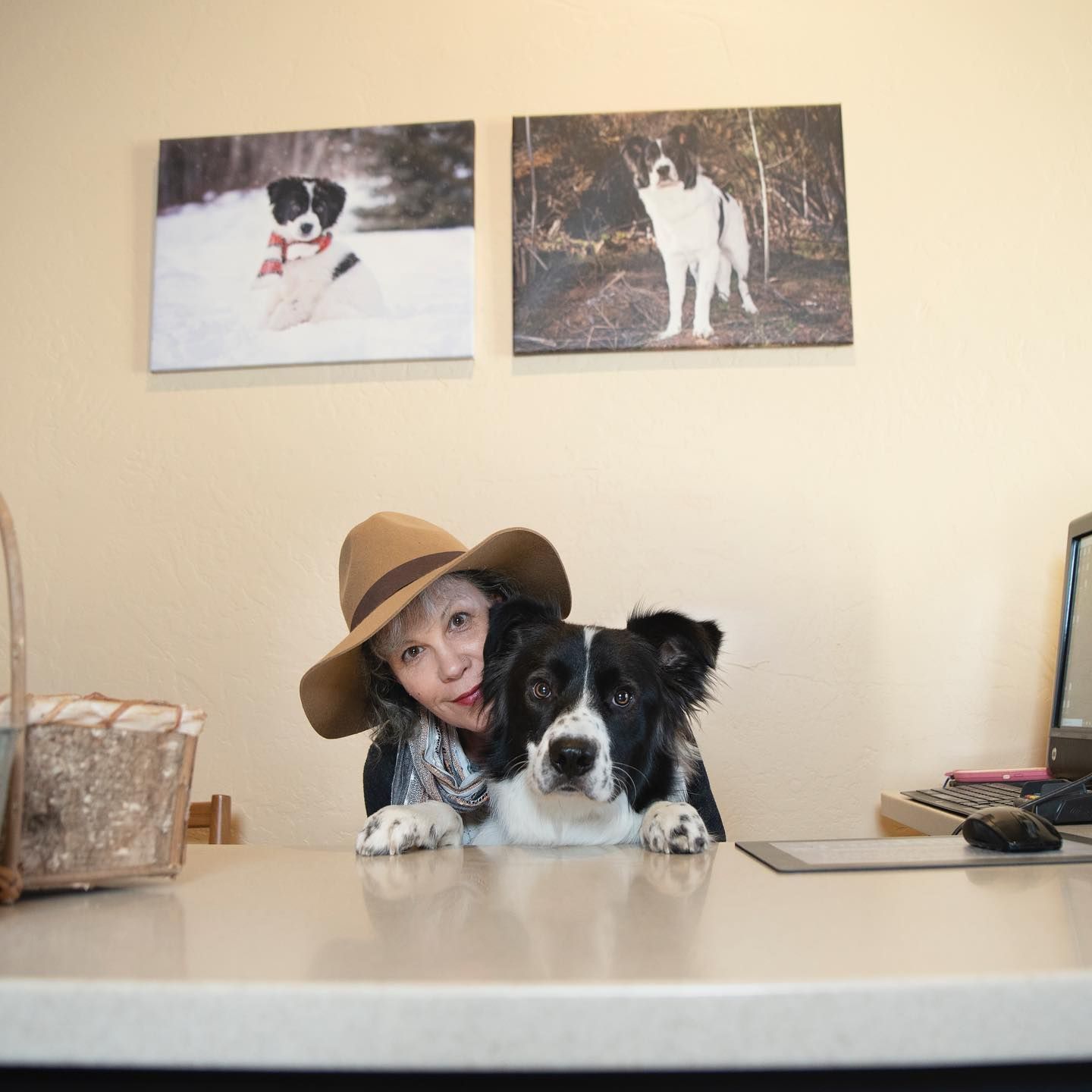 A woman wearing a hat and a black and white dog rest their arms on a table, with two dog portraits hanging on the wall.