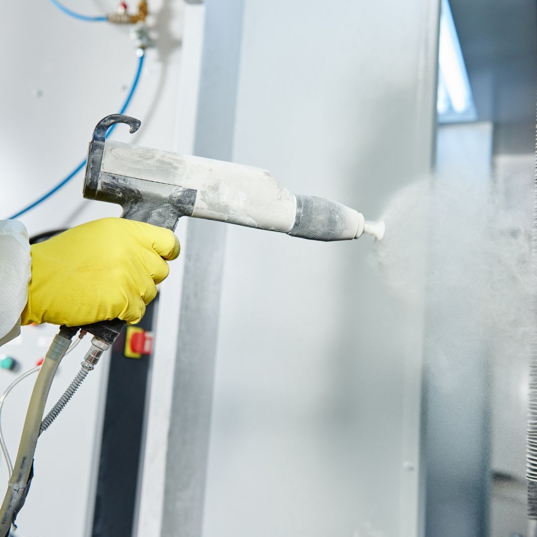 Person in yellow glove sprays powder coating onto a metal surface with a spray gun.