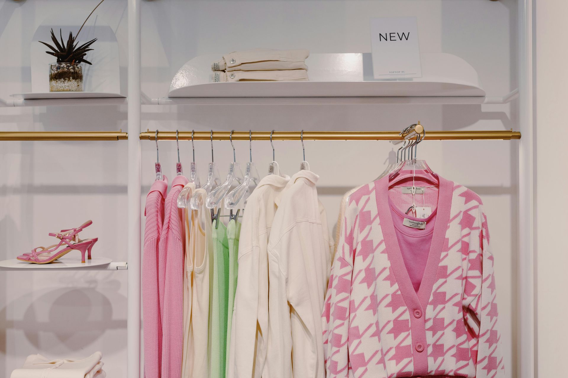 Clothing rack displaying pastel-colored clothes. Pink and white patterned cardigan, shoes, and shelf with decor.