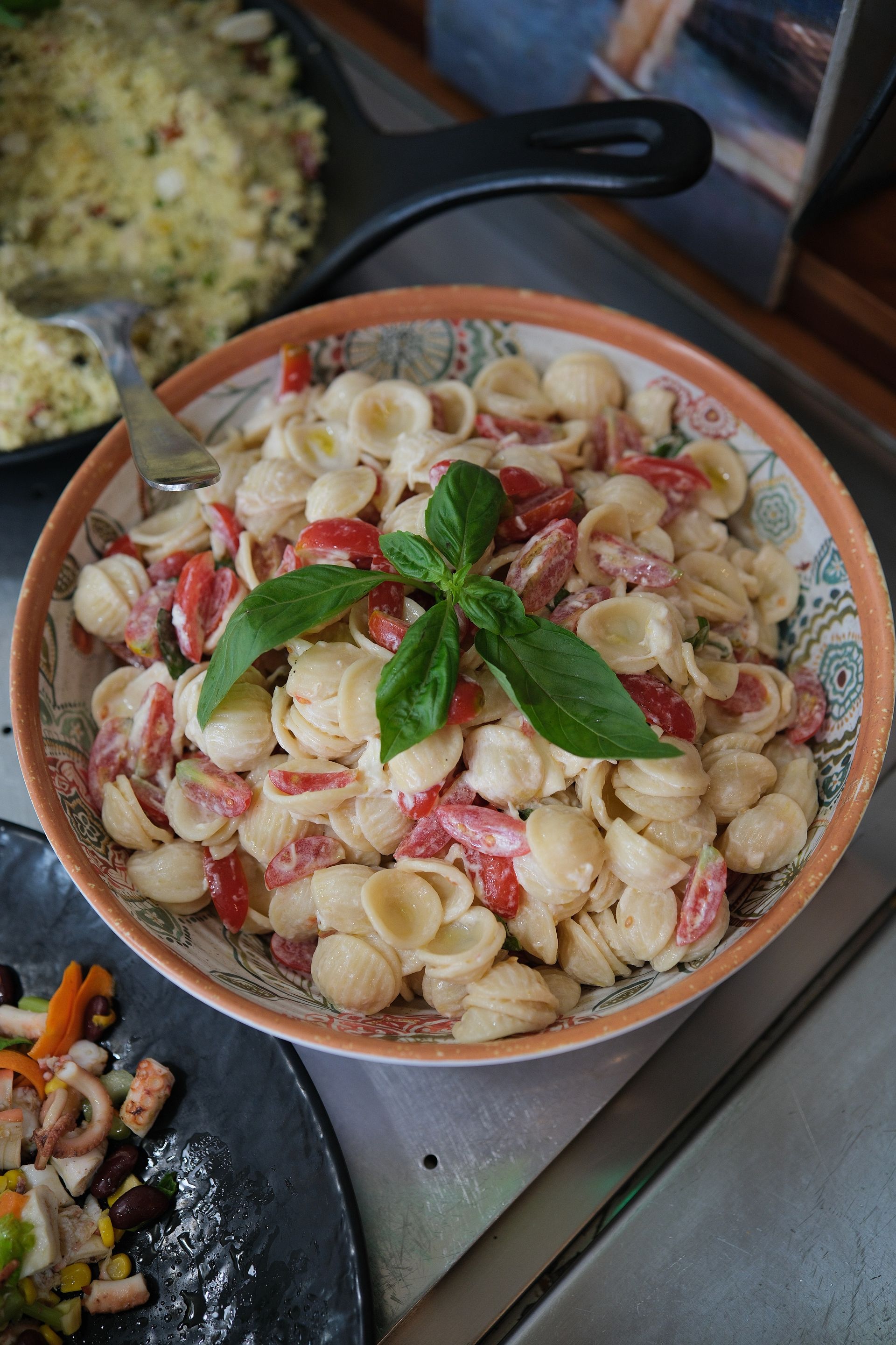Insalata di pasta con pomodori e basilico in una ciotola, circondata da altre pietanze.
