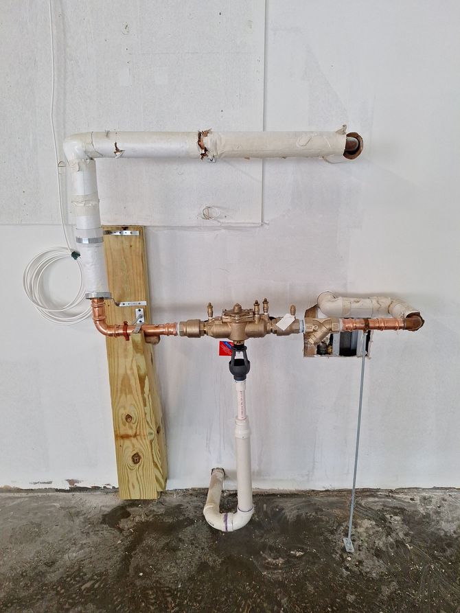 A brass water pressure regulator valve installed on copper piping within a wooden wall frame near white PVC pipes.