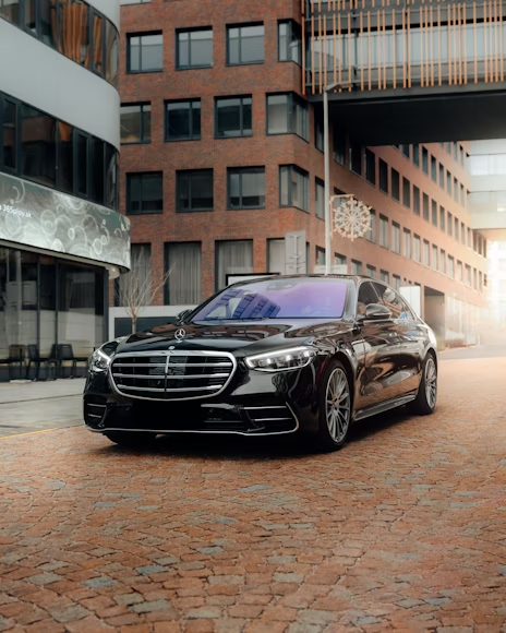 Black Mercedes sedan parked on a brick street between modern buildings, with a bridge overhead.