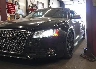 Black Audi car inside a garage, angled shot from the front. Silver grille, head lights on.
