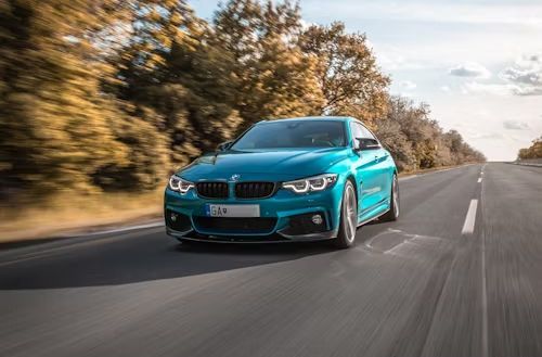 Teal BMW car driving on an asphalt road with trees on the side, sunny day.