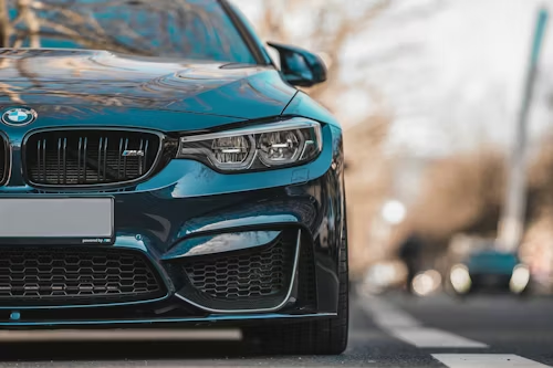 Blue BMW M4 sports car parked on the street with headlights on, blurred background.