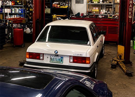 White BMW in a mechanic’s garage, viewed from the rear, with tools and a blue car in the foreground.