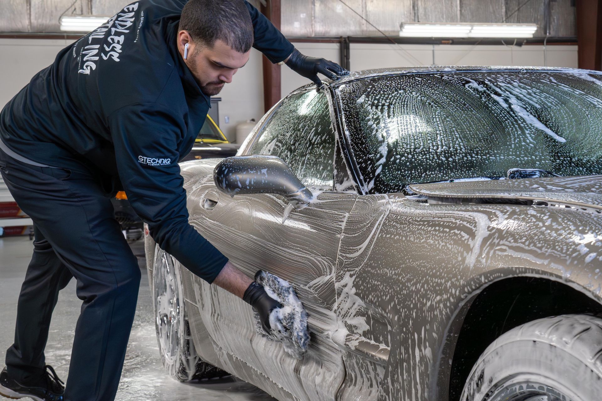Person washing a tan car with soap in a garage, scrubbing the side window and door.