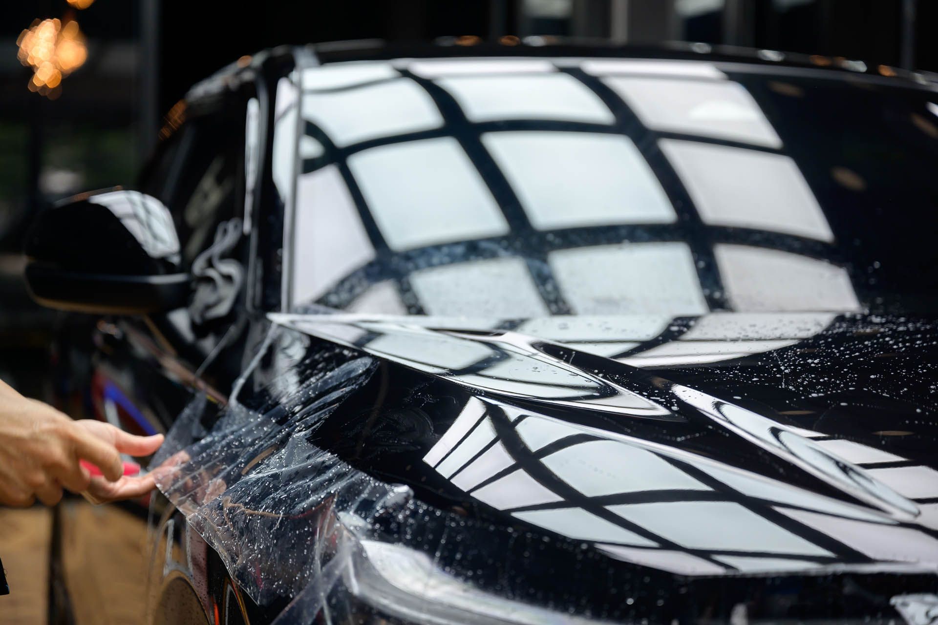 Hand washing a black car with soap suds and water spray under bright window reflections