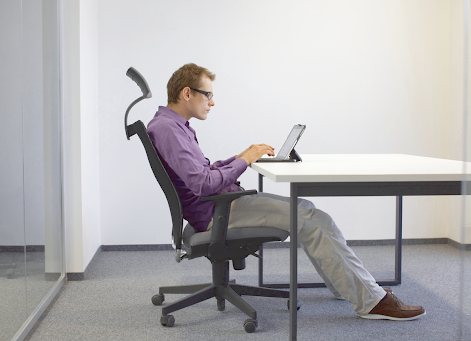 Man in glasses sits at a desk typing on a tablet, leaning back in an office chair.