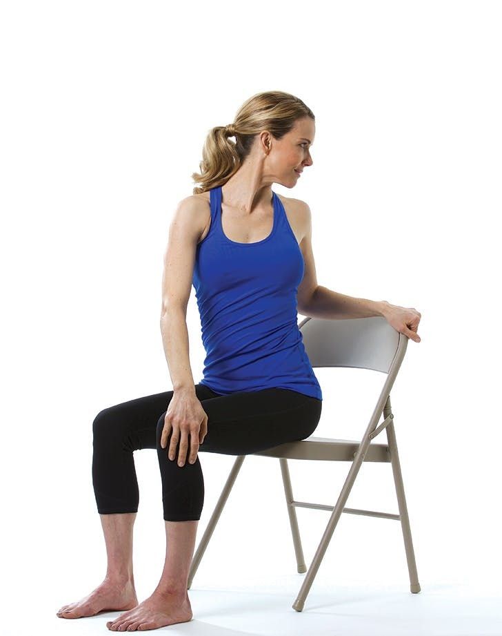 Woman in blue tank top seated, torso twisted, hand on chair back, barefoot.