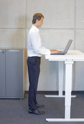 Man in white shirt and dark pants standing at a white adjustable desk, using a laptop in an office.