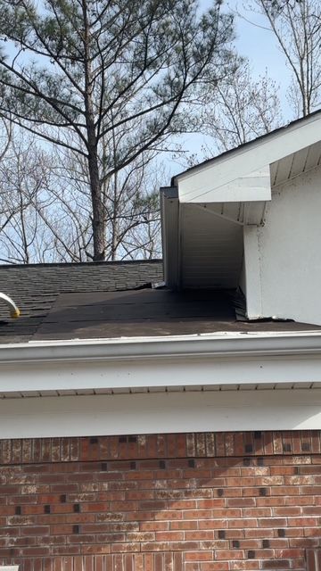 Roof with dark shingles and a white overhang against a brick wall and bare trees. - All American Property Serv