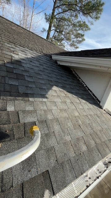 Gray asphalt shingle roof on a house, with a gutter and downspout visible. - All American Property Serv