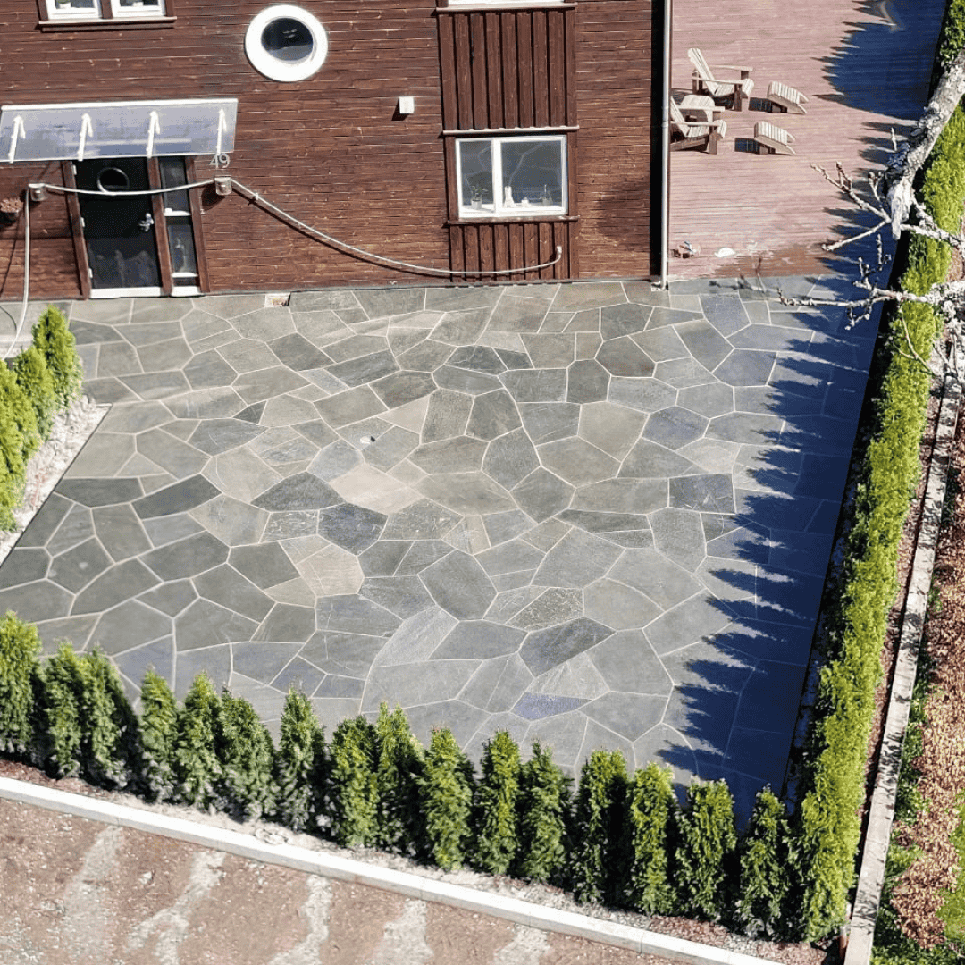 Natursteinsbelagt uteplass med hekk foran bolig.