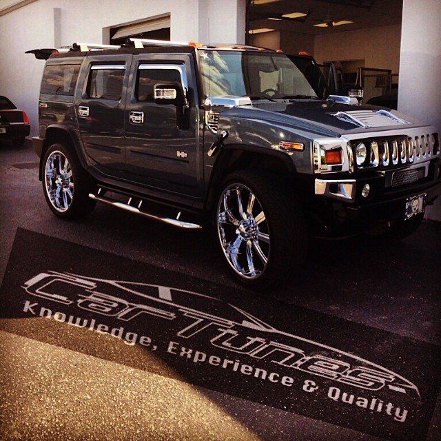 Dark gray Hummer H2 SUV with chrome wheels parked outside a shop; a welcome mat reads 
