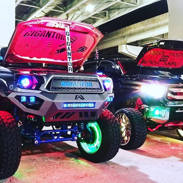 Two modified trucks with open hoods at an auto show, featuring neon lights and custom parts.