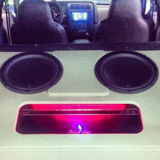 Two large subwoofers and a glowing pink amplifier in a vehicle, forming a face.
