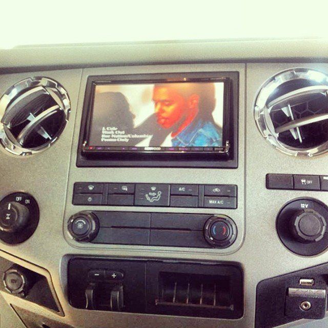 Car dashboard with a screen playing a video of a man in a blue jacket. Silver vents and controls visible.