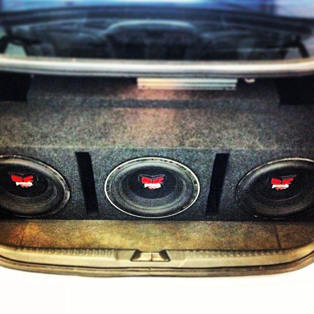 Three subwoofer speakers in a car trunk, mounted in a gray enclosure.