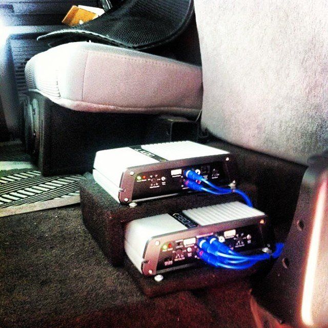 Two car audio amplifiers stacked, with blue cables, under a car seat in a vehicle.