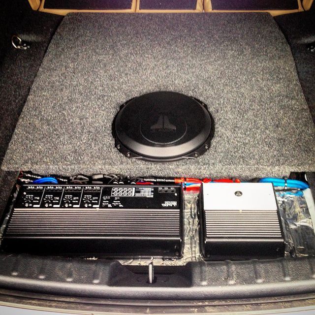 Car trunk with a subwoofer, two amplifiers, and wiring. Gray carpet and black electronics.
