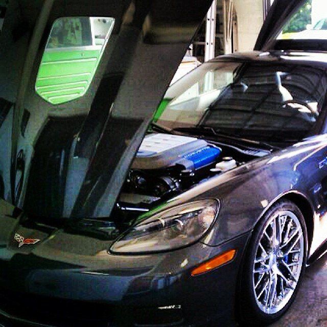 Dark grey Corvette with hood open, exposing a blue engine; front wheel with chrome rim.