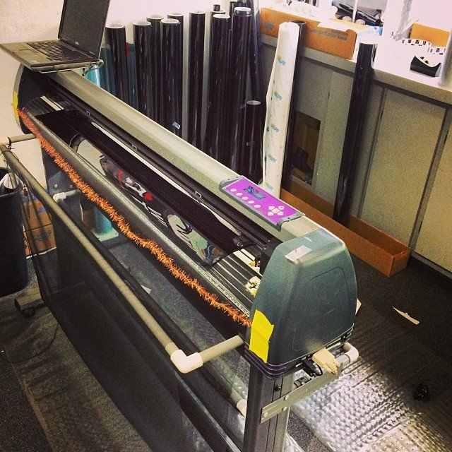 A large format plotter machine with a roll of film cutting a graphic, inside a shop.