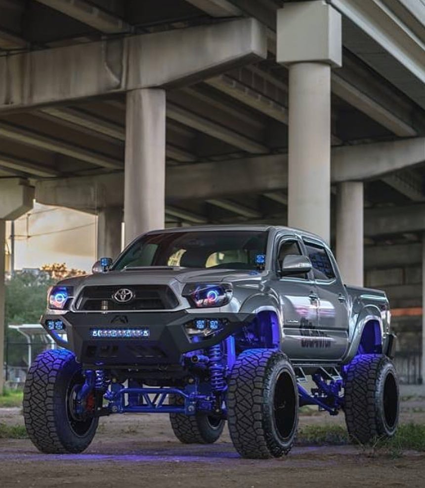 Gray lifted Toyota Tacoma truck with blue underglow lights parked under a concrete bridge.