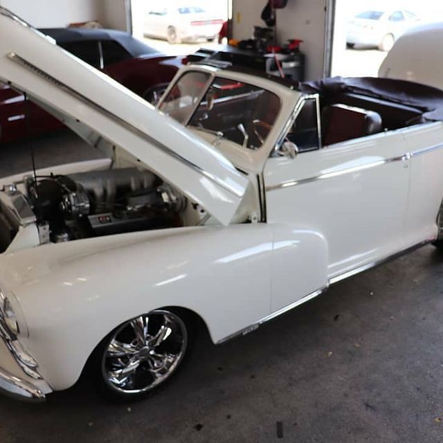 White vintage convertible with hood open, in a garage, shiny chrome wheels.