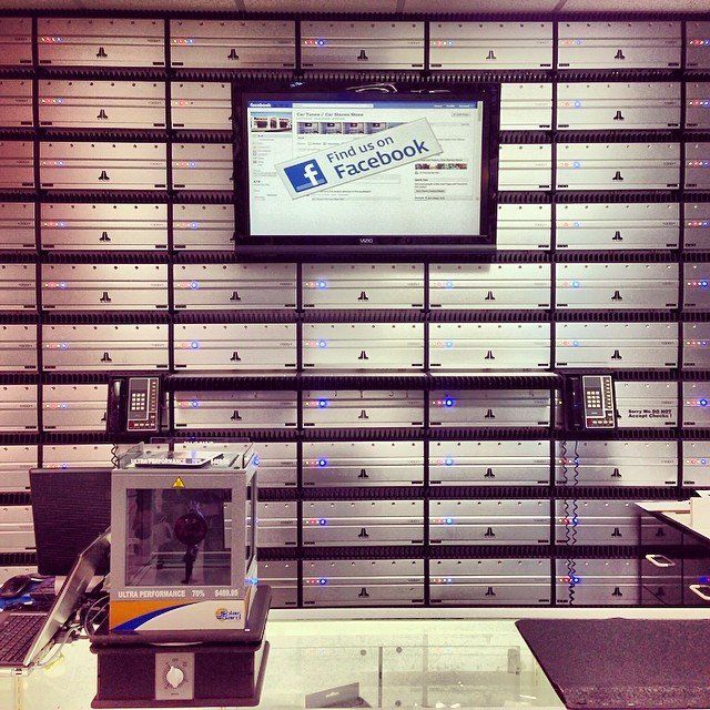 Wall of servers with a monitor displaying a Facebook ad; a 3D printer and keyboard are in the foreground.