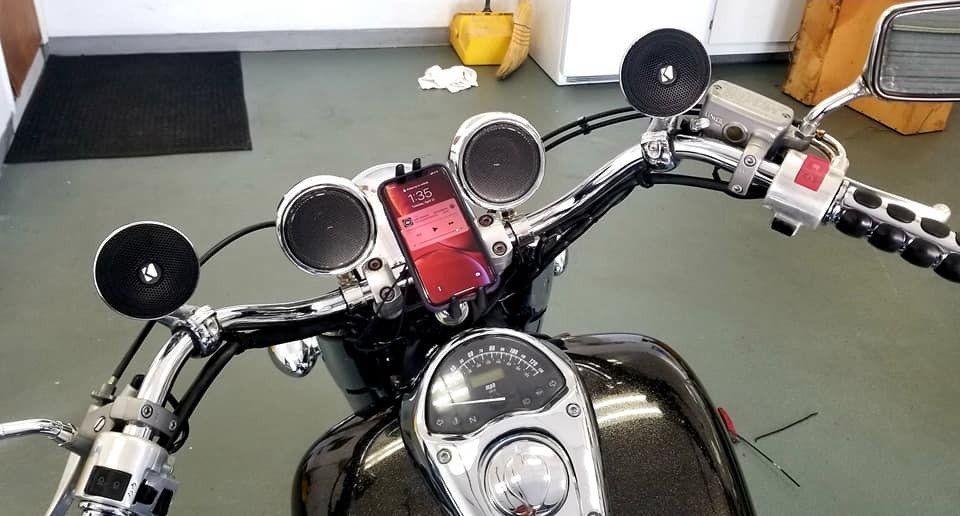 Motorcycle handlebars with phone and speakers mounted; dashboard visible.
