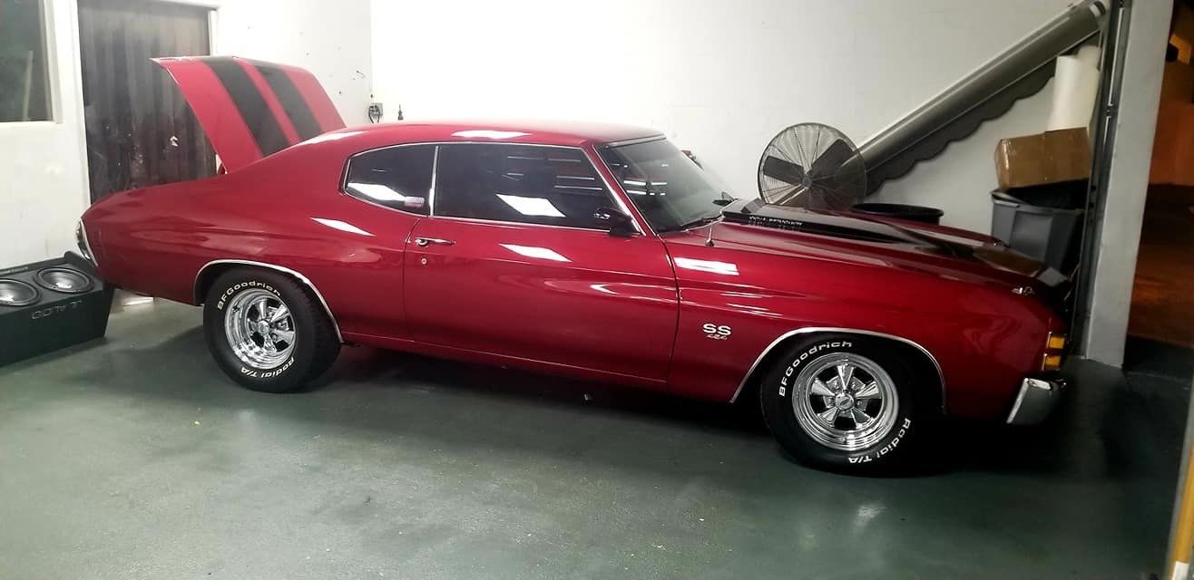 Red classic Chevy Chevelle in a garage. Hood open, black stripes, chrome wheels.