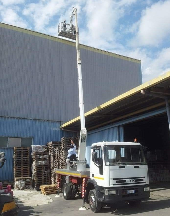 Piattaforma aerea montata su camion accanto a un magazzino, con un operaio sollevato sopra pallet impilati.